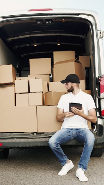 A man wearing a black cap, white t-shirt, and blue jeans is seated on the pavement next to a large open van during a home relocation. The van's cargo area is filled with numerous cardboard boxes of va