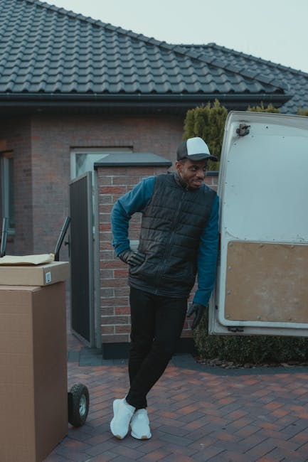 A man wearing a black cap, white t-shirt, and blue jeans is seated on the pavement next to a large open van during a home relocation. The van's cargo area is filled with numerous cardboard boxes of va