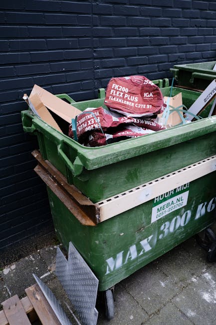 A green waste container filled with several red and black plastic bags labeled 'FLEXIBELBER CZE S1' and 'FK200 FLEXY PLUS,' placed on a paved area outside a building with a dark brick wall. The container is positioned on a wooden pallet, with the top of the bags visible over the open rim, indicating a loading or disposal process related to house removals or clearance. To the right, there is another green waste bin, and a white plastic drainage grid is leaning against the container. The scene suggests preparation for waste removal in the context of a home move or clearing, consistent with professional moving services like those offered by Man with Van Tottenham Green.