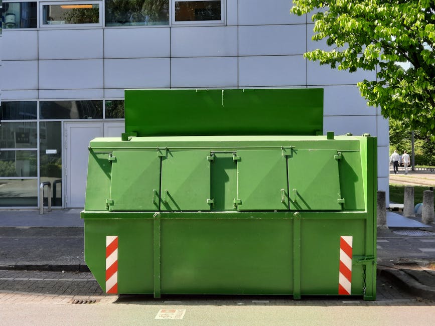 A large green commercial waste skip positioned on the paved sidewalk outside a modern building with a grey tiled facade and large glass windows. The skip is situated beside a white door with a handle, and a metal bike rack is visible nearby. The skip’s lid is closed, and it features red and white reflective safety markings on the lower corners. To the right, a tree with green leaves provides partial shade, and in the background, a few pedestrians are walking along a pathway. The scene suggests a home relocation or rubbish collection process as part of a furniture transport or packing and moving operation, with the skip prepared for bulky waste disposal. Man with Van Tottenham Green occasionally facilitates such waste management and removals services to support comprehensive house moves.