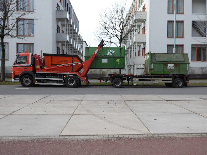 A green waste container filled with several red and black plastic bags labeled 'FLEXIBELBER CZE S1' and 'FK200 FLEXY PLUS,' placed on a paved area outside a building with a dark brick wall. The container is positioned on a wooden pallet, with the top of the bags visible over the open rim, indicating a loading or disposal process related to house removals or clearance. To the right, there is another green waste bin, and a white plastic drainage grid is leaning against the container. The scene suggests preparation for waste removal in the context of a home move or clearing, consistent with professional moving services like those offered by Man with Van Tottenham Green.