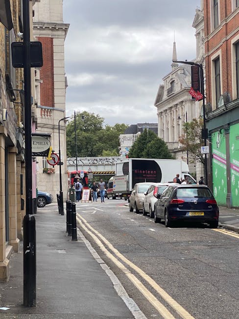 Photograph of a busy street scene on Tottenham High Road during daytime, showing parked cars lining the curb on the right side and a moving van near a loading bay area, where a team of workers is engaged in loading or unloading items for home relocation. The street is paved with asphalt and features double yellow lines along the edge, indicating parking restrictions, with bollards separating the pavement from the road. On the left, shopfronts and signage are visible, including a sign for Emilia, and a pedestrian walkway runs alongside the buildings. In the background, there are historic buildings with classical architecture, a church steeple, and green trees, under a cloudy sky. This scene supports the context of house removals and furniture transport services provided by Man with Van Tottenham Green, illustrating the logistics involved in loading and loading area operations for local commercial and residential moves.