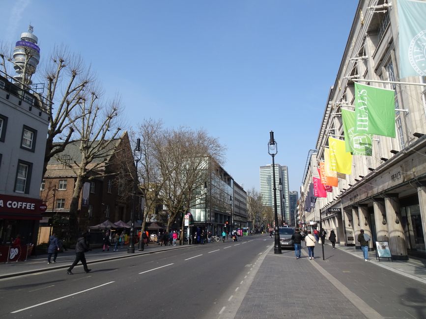 Photograph of a busy street scene on Tottenham High Road during daytime, showing parked cars lining the curb on the right side and a moving van near a loading bay area, where a team of workers is engaged in loading or unloading items for home relocation. The street is paved with asphalt and features double yellow lines along the edge, indicating parking restrictions, with bollards separating the pavement from the road. On the left, shopfronts and signage are visible, including a sign for Emilia, and a pedestrian walkway runs alongside the buildings. In the background, there are historic buildings with classical architecture, a church steeple, and green trees, under a cloudy sky. This scene supports the context of house removals and furniture transport services provided by Man with Van Tottenham Green, illustrating the logistics involved in loading and loading area operations for local commercial and residential moves.