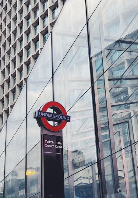 A close-up view of the exterior of a modern glass-fronted building with reflections of surrounding structures. In the foreground, a London Underground sign displaying the line's red, blue, and white colours is mounted on a black pole. Below the sign, a purple-and-white plaque indicates the location as Tottenham Court Road station. The building's glass facade reveals an interior framework and multiple reflections of nearby high-rise buildings, creating a layered urban scene. The lighting suggests daytime with natural light illuminating the glass panels. The setting is in the vicinity of Tottenham Green, where professional removal services such as those provided by Man with Van Tottenham Green may coordinate office relocations for businesses, involving the careful packing, loading, and transport of office furniture and equipment.