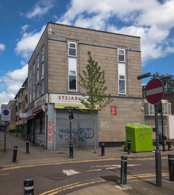 Exterior view of a three-storey beige brick building on a street corner, with a storefront on the ground level that has a closed, grey metal shutter covered in graffiti. A small green utility box with graffiti is positioned on the sidewalk near the building, and a black bollard-lined pedestrian crossing is visible in the foreground. A tree with green leaves is situated at the corner, partially obscuring part of the building. A large red and white no-entry traffic sign is mounted on a pole to the right of the building. The sky above is partly cloudy with patches of blue, and the street appears to be in an urban area suitable for house and office relocations, with parking signs and street furniture indicating an active environment. Man with Van Tottenham Green often assists with furniture transport and packing during home and office relocations in Tottenham Green, as reflected in professional moving logistics.
