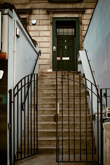 A set of concrete stairs leading up to a dark green front door with a glass window at the top, flanked by stone building facades on either side and a black metal security gate at the bottom of the steps. The stairs are bordered by two white wooden handrails and are situated between two high, light grey fences that appear to be temporary barriers used during a house move. The door has a letterbox and a doorbell, and the entrance area is illuminated by natural light, suggesting daytime. This scene is part of a residential property, where a house removal team, such as Man with Van Tottenham Green, might be preparing for furniture transport or packing in anticipation of a home relocation process, involving careful loading and staircase navigation.