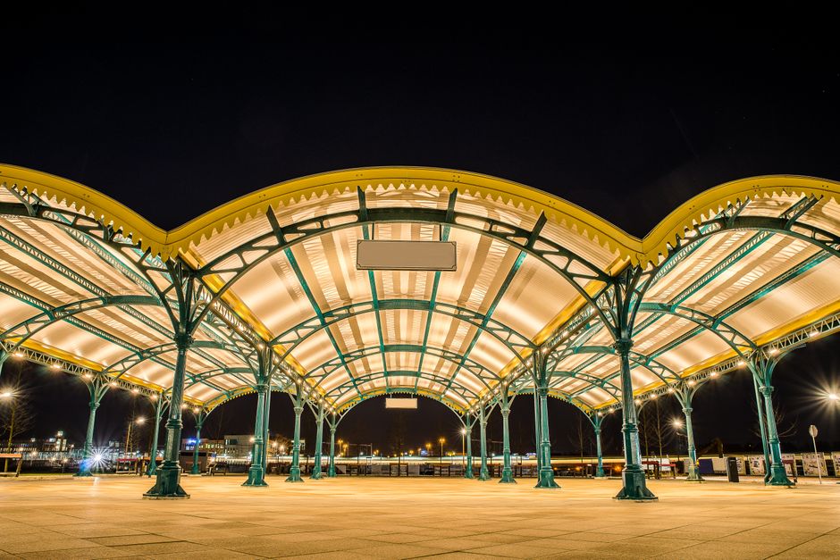 A night-time exterior view of Tottenham Hale station features a large, illuminated canopy supported by green metal columns with decorative curved arches. The canopy's roof is made of translucent panel