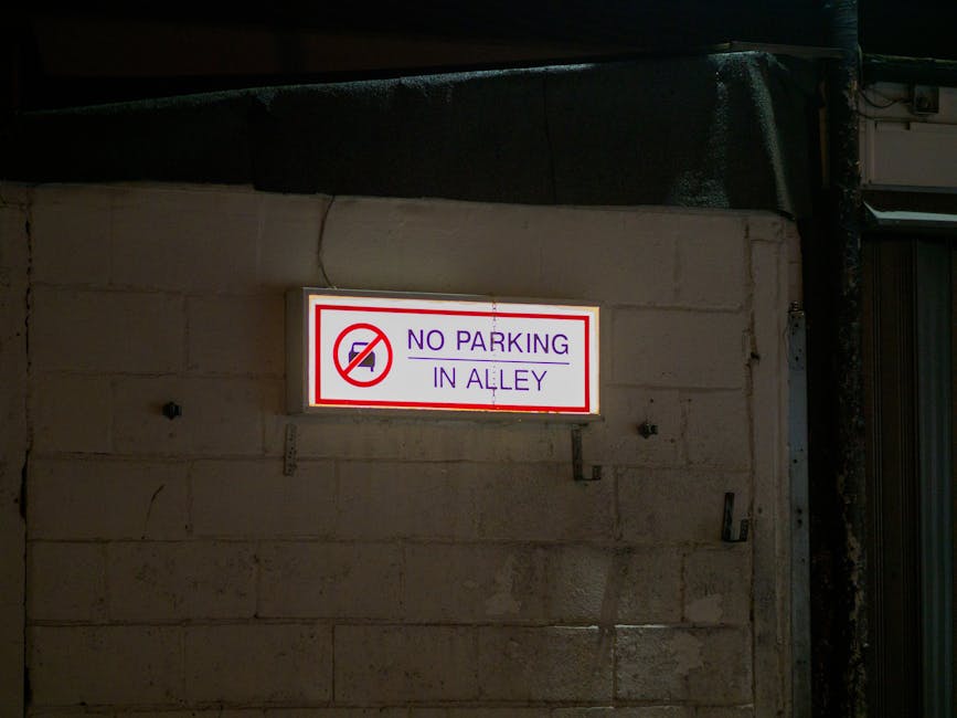 A rectangular white metal sign with bold black text reading 'NO PARKING DAY OR NIGHT' is mounted on a light grey wooden door or shutter. The sign is secured with four metal bolts, one in each corner, and is positioned near the upper middle part of the door. The surrounding environment appears to be part of a building exterior, with horizontal panels and a vertical panel segment visible, indicating a typical setting for residential or commercial property access. The lighting suggests natural daylight, illuminating the sign clearly. This signage is relevant for home relocation or moving logistics, indicating parking restrictions that Movers like Man with Van Tottenham Green need to consider during furniture transport or packing and moving operations in the Tottenham Green area.