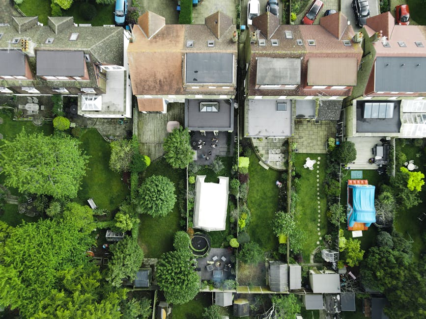 An aerial view of multiple residential backyards in the Tower Gardens Estate area, showing well-maintained gardens with lush green trees, lawns, and a variety of outdoor furniture including a white curved sofa, a black patio table, and several chairs. One backyard features a small trampoline, while another has a blue inflatable pool and a children's play area with an orange slide. Pathways made of paving stones and gravel connect different sections of the gardens, with some areas enclosed by fences or hedges. Visible garage doors, sheds, and garden sheds are present at the rear of some properties. The neighboring houses are detached and semi-detached, with tiled and flat roofs, and are situated close together on a narrow street lined with parked cars. The lighting suggests daytime with overcast skies, capturing the details of the outdoor spaces, part of a home relocation or packing and moving process, typical of house removals services provided by Man with Van Tottenham Green.