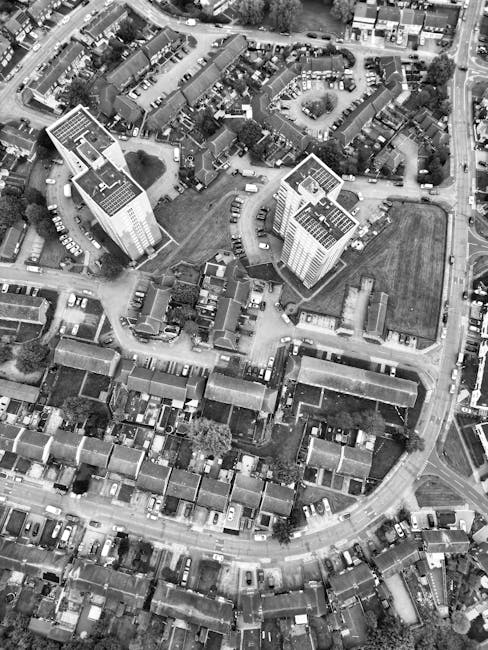 An aerial black-and-white photograph showing a residential neighbourhood with rows of terraced houses and parked cars lining the streets. In the middle of the image, there is a large complex of modern apartment buildings with flat roofs, some featuring rooftop utilities or solar panels. The complex is surrounded by open green spaces and pathways, with parts of the area indicating recent or ongoing building activity, suitable for home relocation planning. In the background, tree-lined roads extend beyond the immediate vicinity, with cars moving along the streets. The scene captures the urban landscape's density, highlighting the variety of housing types and the organized layout typical of residential areas serviced by professional removals and moving companies such as Man with Van Tottenham Green, specializing in furniture transport and home moving logistics.