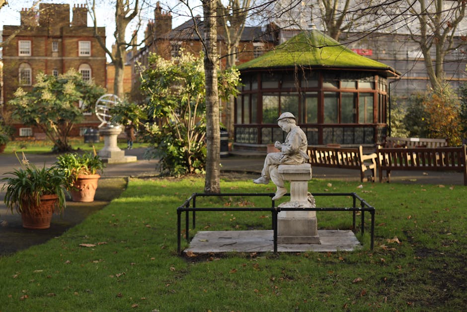 An aerial view of multiple residential backyards in the Tower Gardens Estate area, showing well-maintained gardens with lush green trees, lawns, and a variety of outdoor furniture including a white curved sofa, a black patio table, and several chairs. One backyard features a small trampoline, while another has a blue inflatable pool and a children's play area with an orange slide. Pathways made of paving stones and gravel connect different sections of the gardens, with some areas enclosed by fences or hedges. Visible garage doors, sheds, and garden sheds are present at the rear of some properties. The neighboring houses are detached and semi-detached, with tiled and flat roofs, and are situated close together on a narrow street lined with parked cars. The lighting suggests daytime with overcast skies, capturing the details of the outdoor spaces, part of a home relocation or packing and moving process, typical of house removals services provided by Man with Van Tottenham Green.