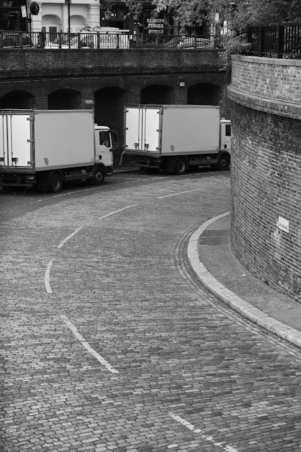 Two white moving trucks parked on a curved brick-paved street beneath a brick bridge with arches and a black metal railing. The trucks are positioned side by side, facing away from the camera, with the rear doors closed. The setting appears to be an urban area, with a high brick wall on the right side and some greenery visible above the railing. The scene captures the loading process typical of home relocation or furniture transport, with no specific household items or furniture visible in this phase of the move. Natural daylight illuminates the scene, highlighting the cobbled street and the vehicles' clean, white exteriors. This image may represent the logistics aspect of a house removal service, such as [COMPANY_NAME], preparing for a transfer of personal belongings in Tottenham Green as part of their same-day removals service.