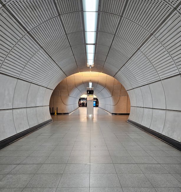 A wide indoor tunnel with smooth, curved metallic walls and a perforated ceiling, illuminated by centered linear lighting. The floor is tiled with large, light grey tiles, and the space appears to be part of a modern transportation hub or station, with no visible furniture or moving equipment. The tunnel extends forward towards a distant, bright exit or opening, creating a symmetrical perspective. In the context of house removals or relocation, this environment can represent a transit point where furniture and boxes are transported through or processed during a home relocation, with Man with Van Tottenham Green handling the loading process in similar indoor environments.